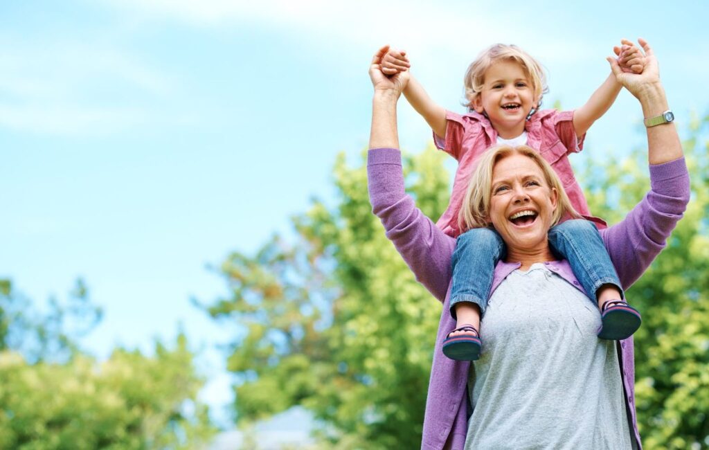 Woman is smiling and holding granddaughter on her shoulders in outdoor space.